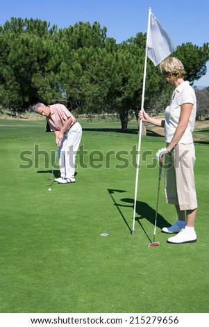 Mature couple standing on putting green, man playing shot, woman holding golf flag, watching