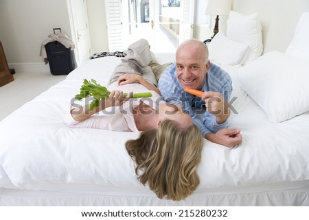Mature couple lying on bed holding carrot and celery sticks, smiling, elevated view
