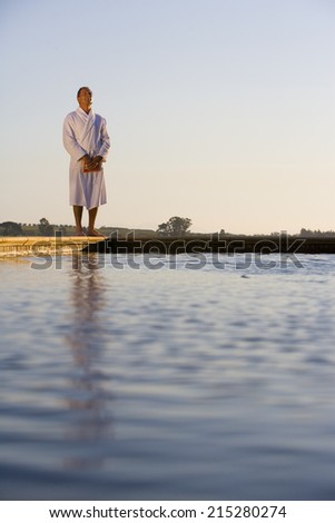 Senior man wearing white bath robe, standing outdoors by swimming pool, eyes closed