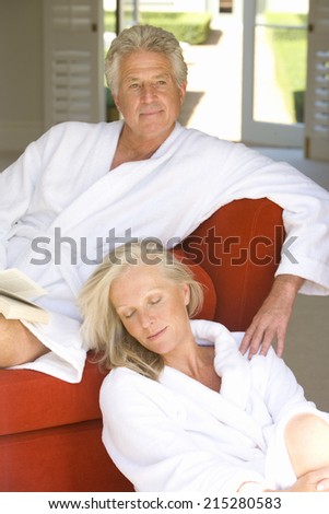 Mature couple wearing white bath robes, woman sitting on floor by man with book on sofa