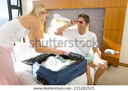 Young couple packing suitcase on bed, man wearing sunglasses, smiling at woman