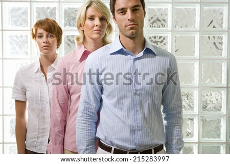 Two businesswomen and man standing in line by glass block wall, portrait