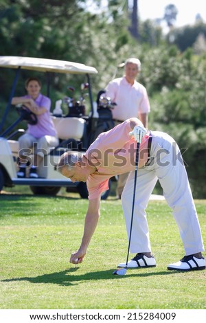Three mature adults playing golf, mature man in pink polo shirt preparing to tee off, mature couple watching from golf buggy in background