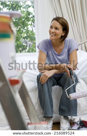 Young woman on sofa with paint roller, paint pot on ladder in foreground (differential focus)