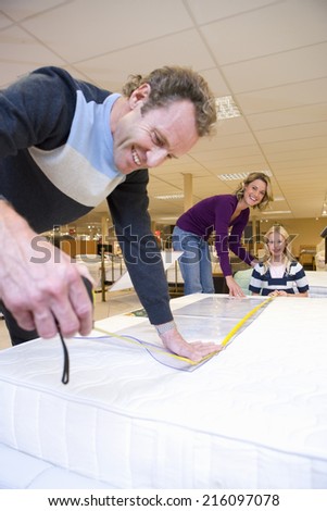 Man measuring bed in furniture shop, family in background, smiling