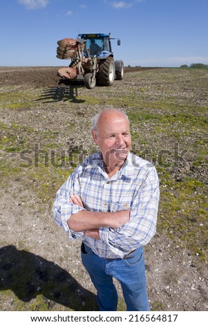 Smiling farmer standing with arms crossed in field with tractor and plough in background