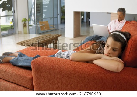 Girl lying down on a sofa, father working on the laptop.