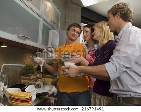 Young man washing dishes and his friends standing behind him with stacks of dishes