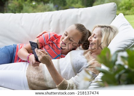 Smiling grandmother and granddaughter laying on outdoor sofa with digital tablet