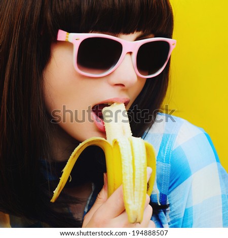 Portrait of beautiful sexy girl in a blue shirt and pink glasses on an orange wall with a banana in his hand to his lips