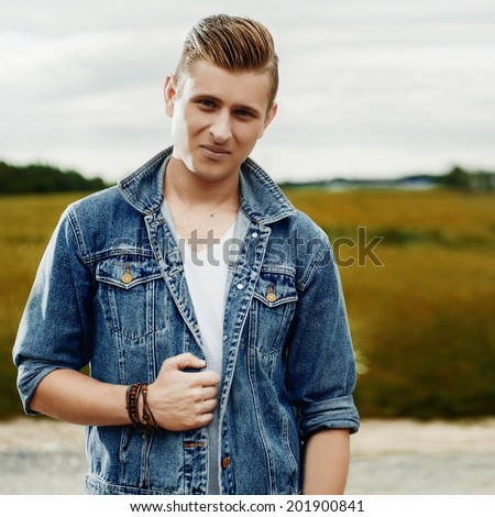 Portrait of a young man in a denim jacket and fashionable hairstyle standing in the field