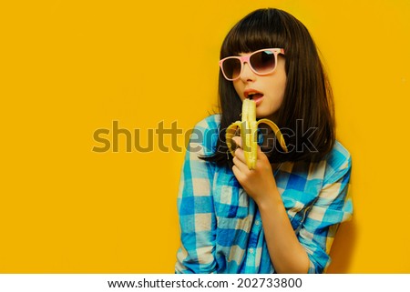 portrait of a girl in a blue shirt on orange background wearing sunglasses. girl eating a banana