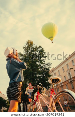 portrait of a beautiful girl with a bicycle in a hat looking at flying the balloon in the city outdoor