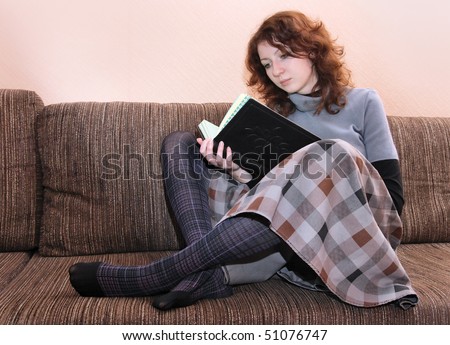 Girl holding up a book and reading intently. Young woman with curly red hair sitting on the sofa