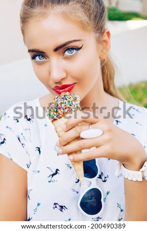 Close up outdoor fashion portrait of young sexy blonde with big blue eyes and full red lips , eating ice cream in city park, wearing cute white outfit.