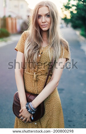 Outdoor fashion portrait of young pretty blonde girl wearing stylish retro vintage yellow dress and perforated leather bag. Posing at old european town.