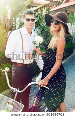 Young handsome man present flower to his girlfriend at first date. Posing outdoor in city garden with retro bike, wearing stylish vintage outfit and sunglasses.