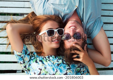 Young pretty blonde girl and her handsome boyfriend laying outdoor in white chubby chic wooden floor, hugs and enjoy the sun and romantic time together.