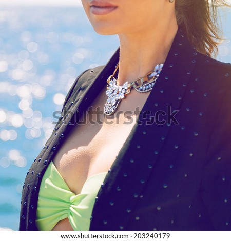 Close up fashion image of woman posing near sea with big sparkling diamond necklace neon bikini and black jacket.