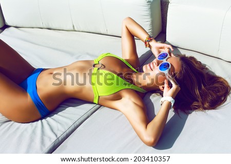 Hight fashion portrait of young tan fit sexy model posing in hot summer day in big white beach sofa, wearing stylish bikini and sunglasses.