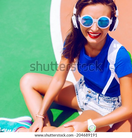 Young pretty happy sportive fit woman sitting in sports ground at retro jeans shorts bright t-shirt and sunglasses, white backpack and earphones, enjoy summer hot day in sports ground.