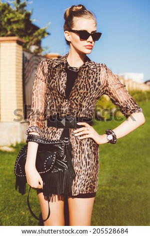 Outdoor fashion portrait of young beautiful stylish woman wearing mini printed dress casual black leather bag with spikes, and retro cat eye sunglasses.