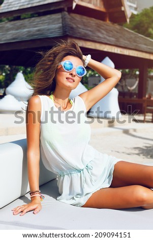 Beautiful stylish girl resting ant enjoy summer at luxury resort on her vacation, sitting at big white sofa in light dress and blur sunglasses.