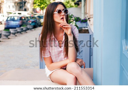 Fashion portrait of young model with long straight brunette hair posing at the street wearing hipster round sunglasses and plaid shirt.