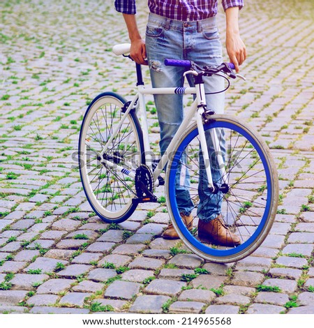 Close up fashion image of stylish man in denim pants and plaid shirt posing with fix bike in old europe city.