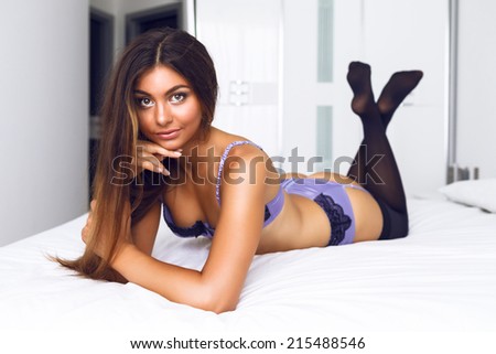 Sexy brunette tan girl with perfect skin and body boxing at big white bedroom on trendy purple silk cab are styled lingerie, relaxing and looking on camera.