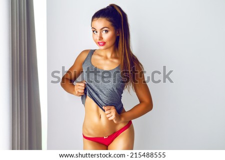 Indoor fashion portrait of sportive girl with perfect fitness tanned body posing at her bedroom on white background, have bright make up and long brunette hairs.