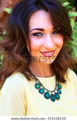 Close up fashion portrait of beautiful woman with full lips amazing smile and bright cat eye make up look, wearing soft yellow blouse and big stylish diamond necklace. Outdoors.