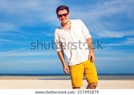Outdoor fashion portrait of handsome man posing at amazing tropical beach, in nice sunny day, beautiful view on blue sky and ocean, wearing casual yellow sorts classic white shirt and sunglasses.