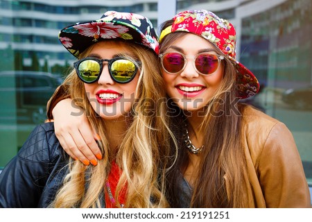 Close up fashion portrait of two pretty girls , wearing leather jackets swag hats and mirrored sunglasses, having blonde and brunette hairs and bright lips. Best friends positive portrait.