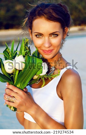 Close up fashion portrait of beautiful bride with fresh natural make up and simple white top, posing with exotic lotus bouquet at sunset on the beach.