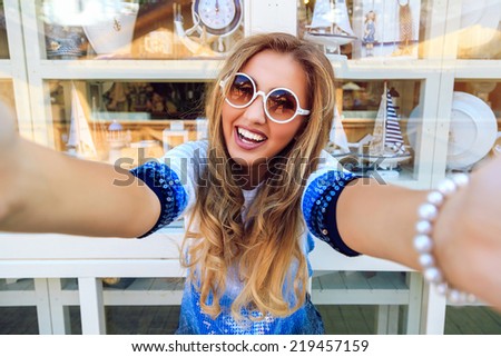 Happy smiling girl trying to take your camera, funny playful image of laughing woman posing near souvenir window shopping bright stylish sweater and sunglasses.