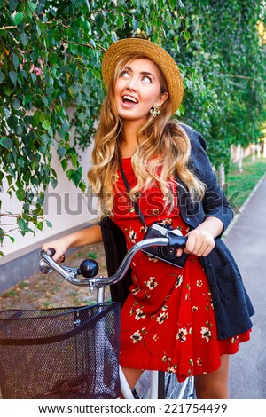 Happy smiling woman having fun and surprised playful emotions, walking alone with stylish retro bike in city park, wearing red dress warm sweater and vintage straw hat, have curled long blonde hairs.