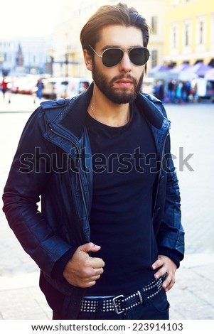Young handsome stylish biker man, posing on the street, wearing stylish black leather jacket and classic trendy sunglasses. Evening fall soft sunlight.