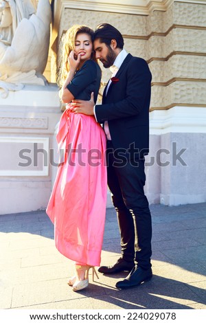 Fashion outdoor portrait of young stylish elegant sexy couple posing at the street. Wearing elegant glamour evening suit and maxi dress, bright sunny colors. First romantic date.