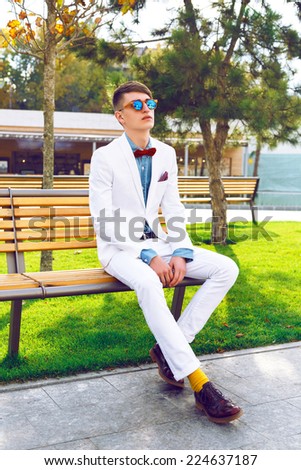 Fashion lifestyle portrait of young hipster man posing in city park in nice sunny day, sitting on the bench at stylish trendy white suit, denim shirt, bow tie, and sunglasses. Bright vintage clothes.