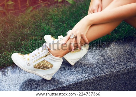 Close up fashion image of woman legs , wearing golden pumped sneakers, and big diamond ring, sitting on the grass, bright colors.