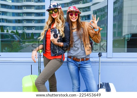 Funny lifestyle portrait of pretty best friends girls having fun before their trip, posing with  baggage near airport, wearing bright casual sportive clothes and sunglasses, ready for new emotions.