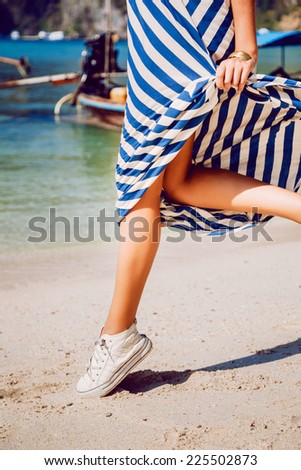 Outdoor fashion image of woman legs jumping at exotic beach in the front of amazing clear ocean, wearing stylish dress and sneakers.