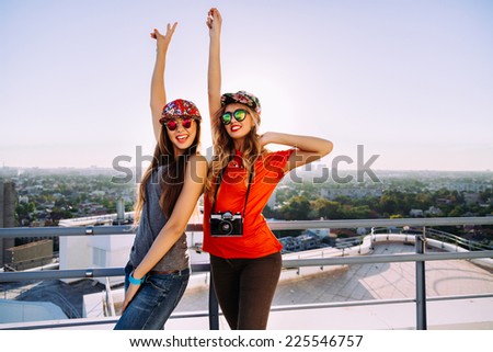 Outdoor lifestyle portrait of two pretty stylish best friends posing on the roof with amazing view to the city, put their hands to the air screaming laughing going crazy and enjoy their freedom.