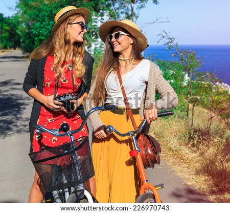 Pretty young smiling girls walking near sea side at nice sunny day, wearing vintage outfits, sunglasses, and hats, taking pictures on retro camera and riding bright hipster bicycles.