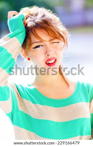 Outdoor portrait of pretty young teen girl making funny faces and winks to you, put her hand to her had imitating headache. Bright colors.