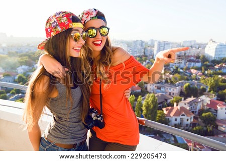 Two pretty girl posing on the roof, hugs and show something, have surprised positive emotions, sisters wearing bright stylish hipster swag clothes and sunglasses.