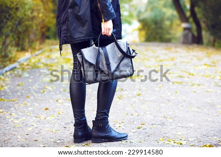 Fashion outdoor image of stylish woman wearing leather pants, warm parka and trendy boots, holding grey bag. Posing at the city park in fall autumn day in the front of yellow leaves.