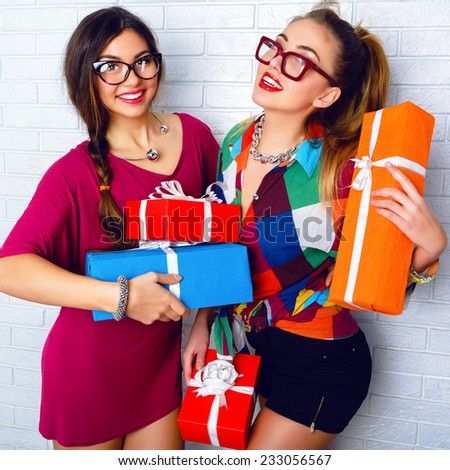 Indoor portrait of two happy best friends sisters girls, ready for holiday party, holding vivd bright gifts and presents, wearing bright clothes and hipster glasses. Urban white background.