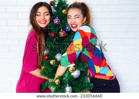 Two pretty best fiends girls posing near decorated Christmas tree, wearing bright party clothes, ready for celebration, smiling and have positive emotions before holidays. Cute indoor family portrait.